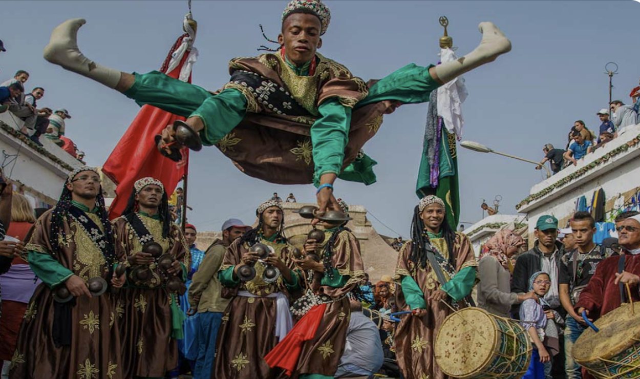 A Gnaoua dancer leaps mid-air above musicians playing krakebs and drums during a street celebration in Morocco