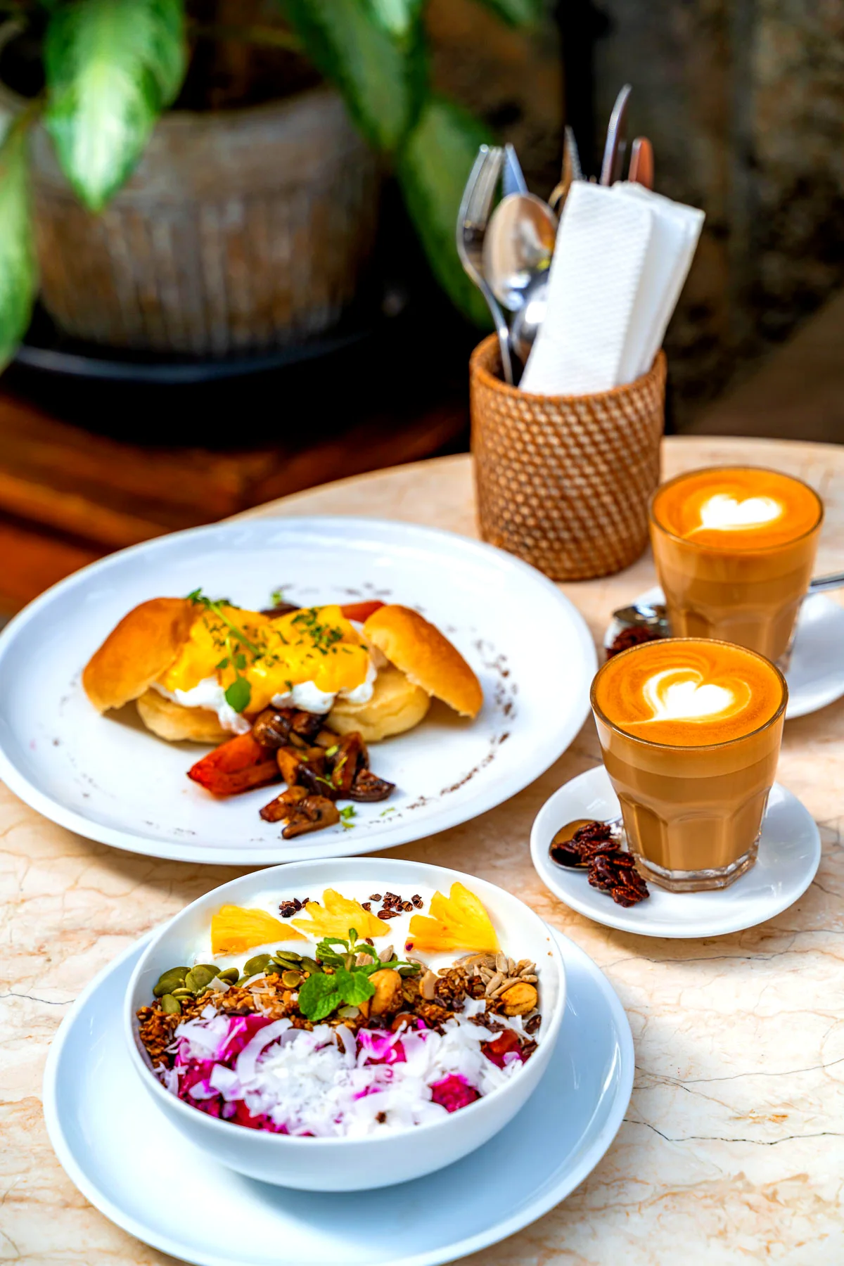 Two lattes with heart latte art beside eggs Benedict and a fruit-and-granola bowl on a marble table at a café in Koh Phangan